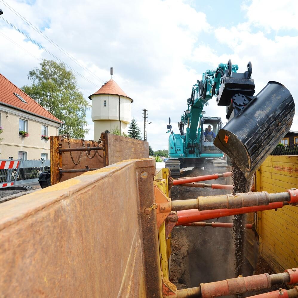 Bagger in einem tiefen, gelben Grabenverbau in einem Dorf mit historischem Gebäude.