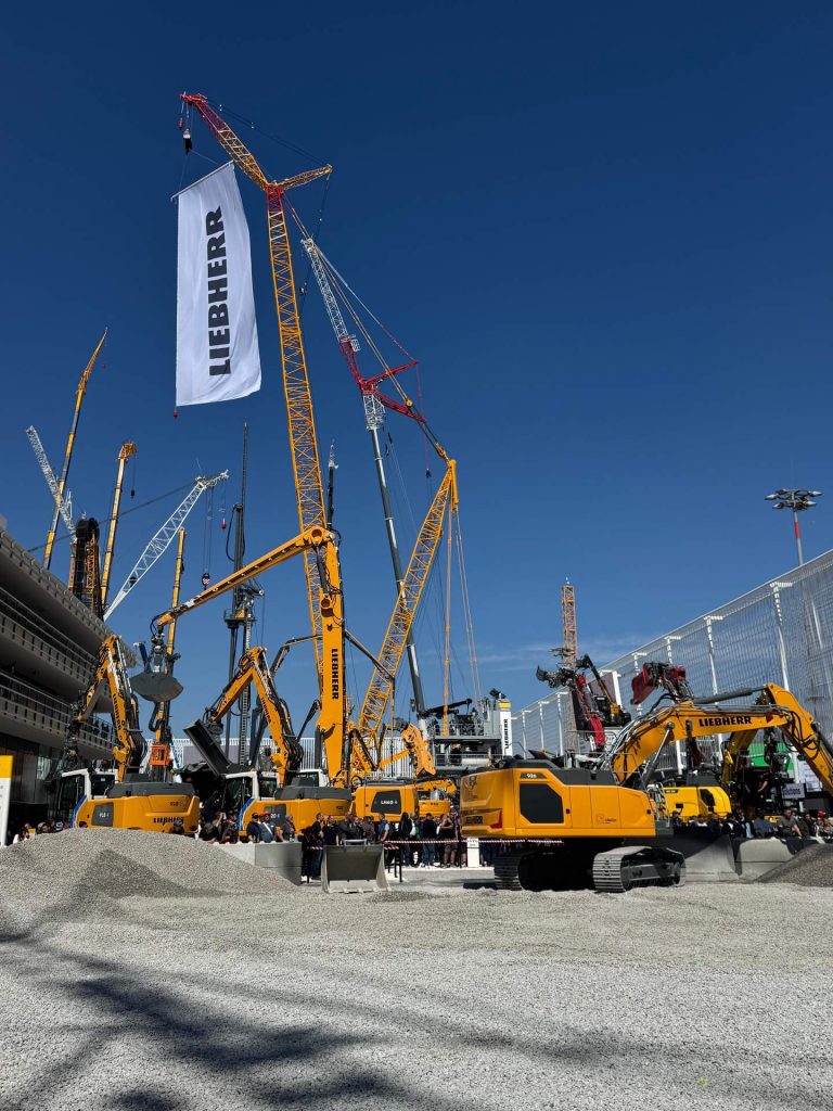 Ein großer Stand der Marke Liebherr auf der Bauma im Freien. Verschiedene Kräne und Bagger in Gelb sind auf einem Schotterplatz ausgestellt. Ein großes weißes Banner mit dem Liebherr-Logo hängt an einem der Kräne.