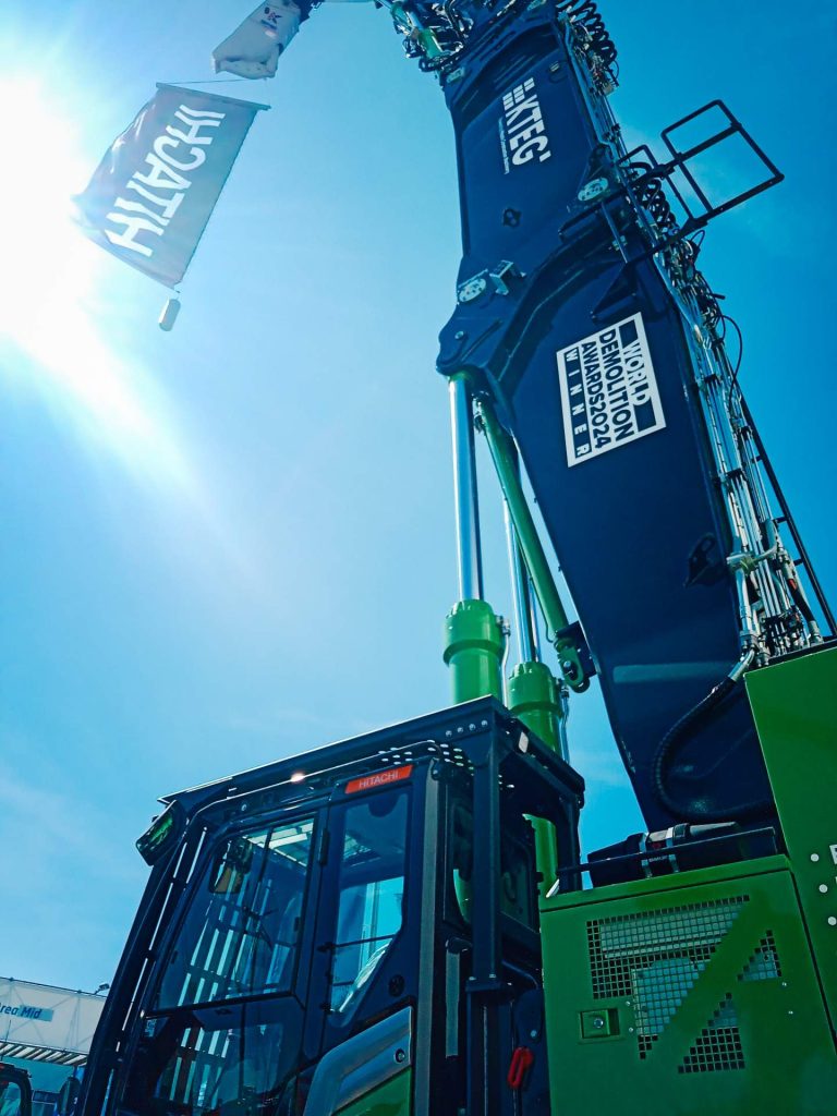 Blick von unten auf einen großen grünen Hitachi-Bagger mit einem großen Banner, auf dem die Aufschrift "World Demolition Awards 2024 Winner" steht.