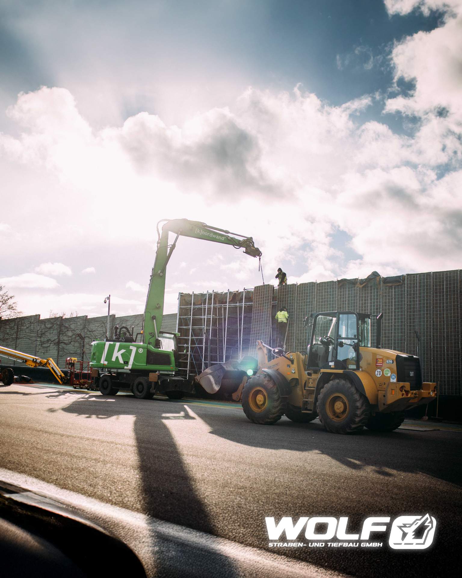 Großer grüner Bagger und gelber Radlader arbeiten zusammen beim Bewegen von Materialien an einer hohen Lärmschutzwand.