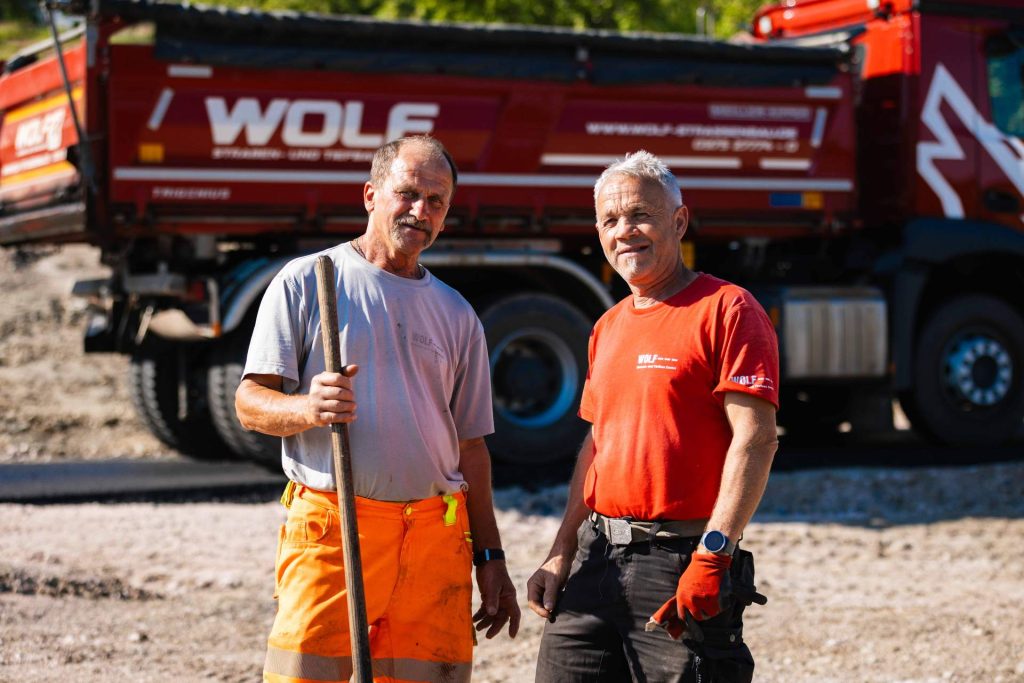 Zwei zufriedene Mitarbeiter von Wolf Straßenbau posieren vor ihrem roten LKW, der beim Transport von Baumaterialien im Kurpark Schlema eingesetzt wurde.
