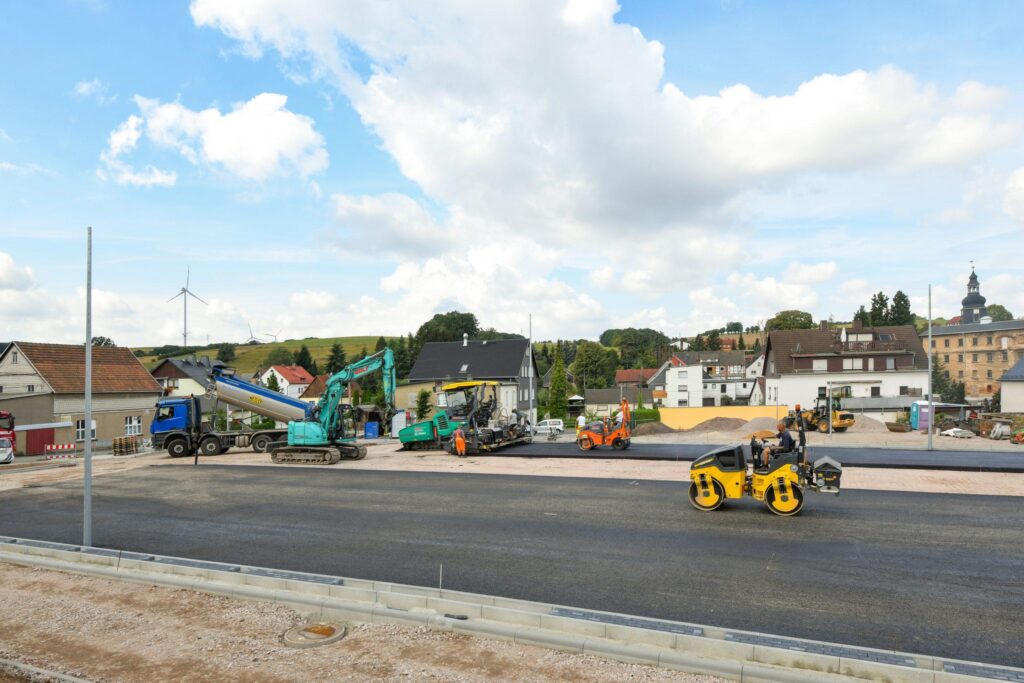 Baustelle mit vielen Maschinen (Fertiger, Walze, LKW); frischer Asphalt.
