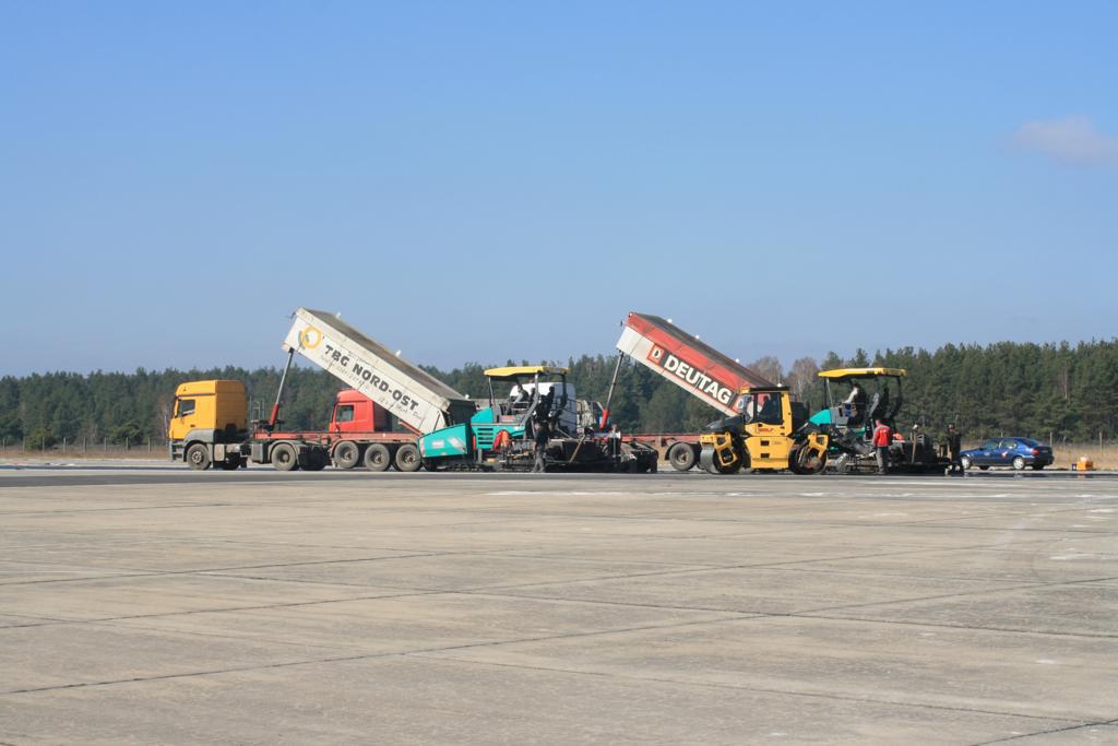 Der Einsatz modernster Asphalt-Baumaschinen: hier sind mehrere Spezialfahrzeuge beim Materialtransport und -einbau zu sehen, vor blauem Himmel.