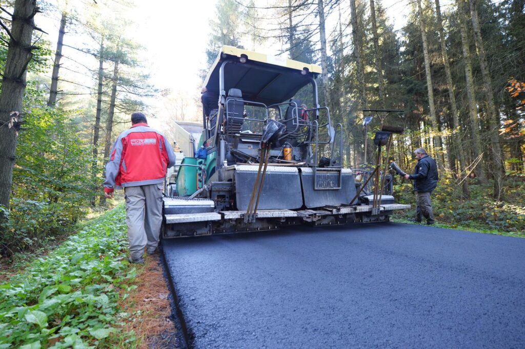 Asphaltfertiger verlegt die Deckschicht; Mitarbeiter kontrolliert den Rand.