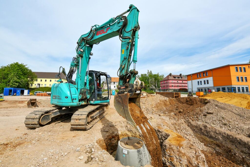 Bagger hebt in Mülsen Graben für Rohre/Kabel aus; alte und neue Gebäude im Hintergrund.