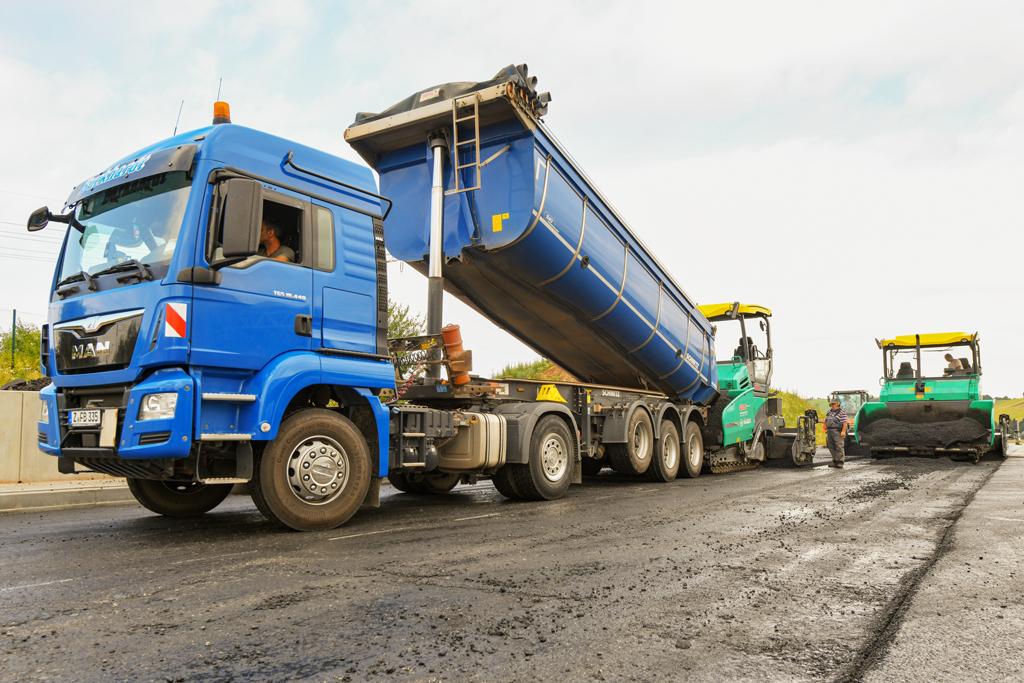 Asphaltierung: LKW-Kipper liefert frisches Asphaltmischgut an Straßenfertiger auf der Baustelle.