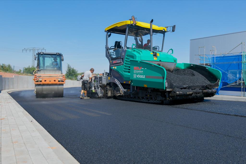 Walze und Straßenfertiger arbeiten zusammen beim zweilagigen Asphalteinbau zur Flächenversiegelung.