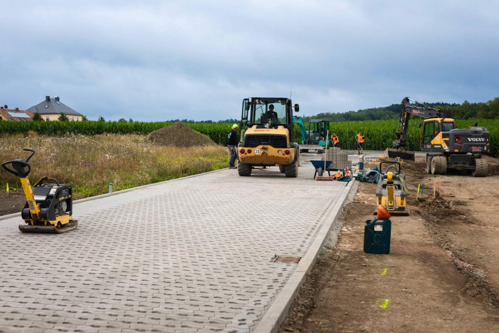 Baustelle mit zwei Bauarbeitern und verschiedenen Baumaschinen, darunter eine gelbe Rüttelplatte, ein gelber Walzenlader und ein Bagger. Ein gepflasterter Weg führt von vorne ins Bild. Im Hintergrund sind Maisfelder, Bäume und Häuser zu sehen.