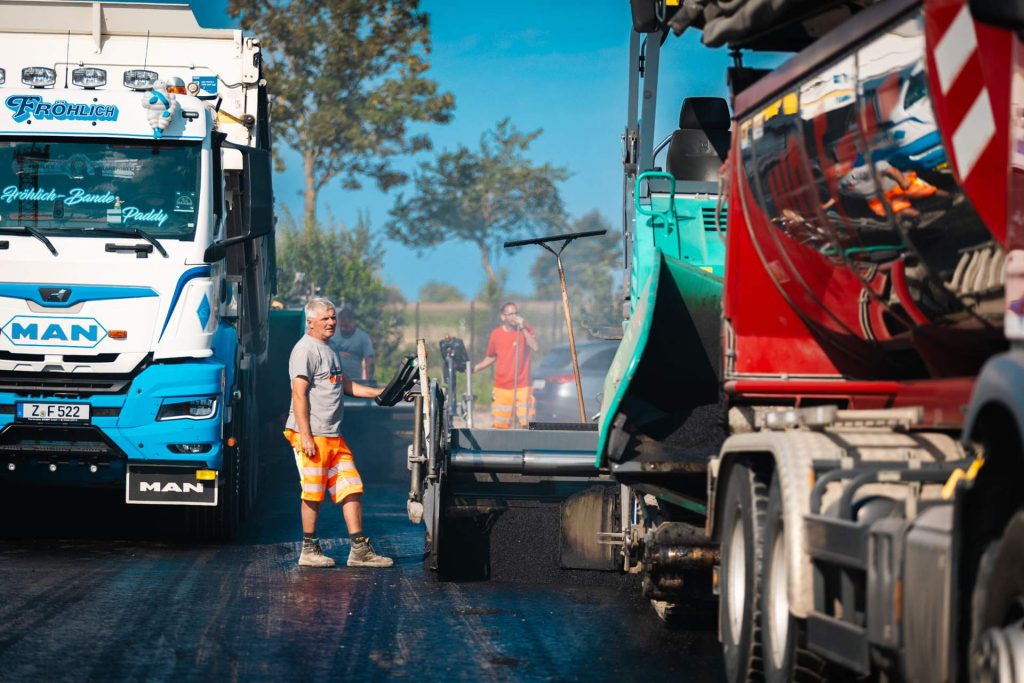 Asphaltarbeiten auf einer Straße mit Arbeitern und Baufahrzeugen, darunter ein MAN-Lkw und ein Asphaltfertiger.