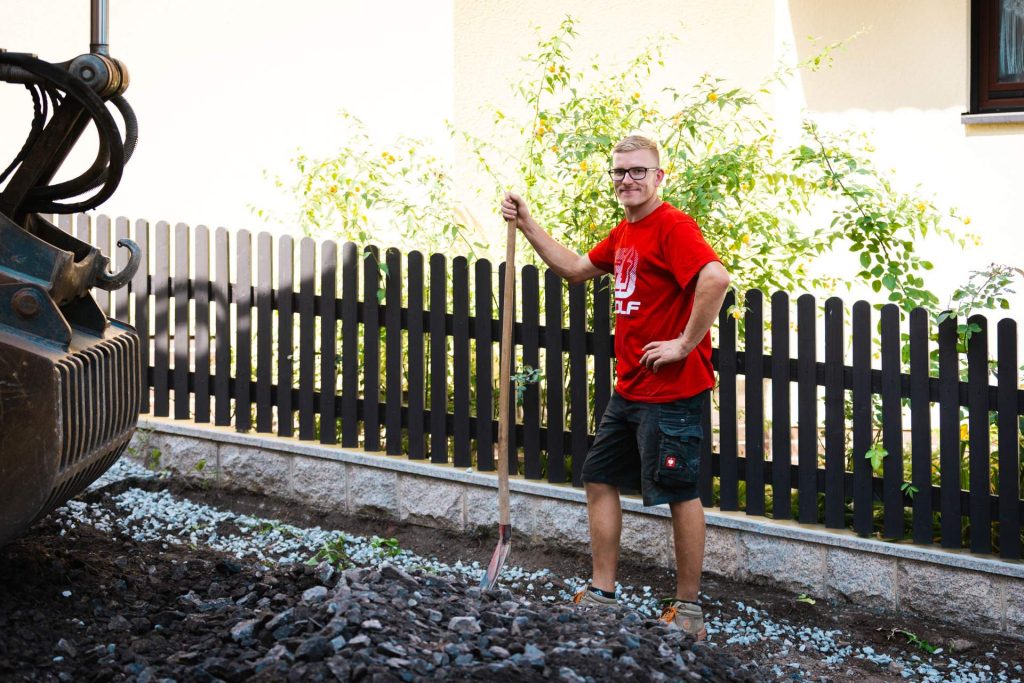 Ein Mitarbeiter posiert mit einer Schaufel vor einer Baustelle. Im Hintergrund ist ein Teil eines Baggers und die Fassade eines Wohnhauses zu sehen.
