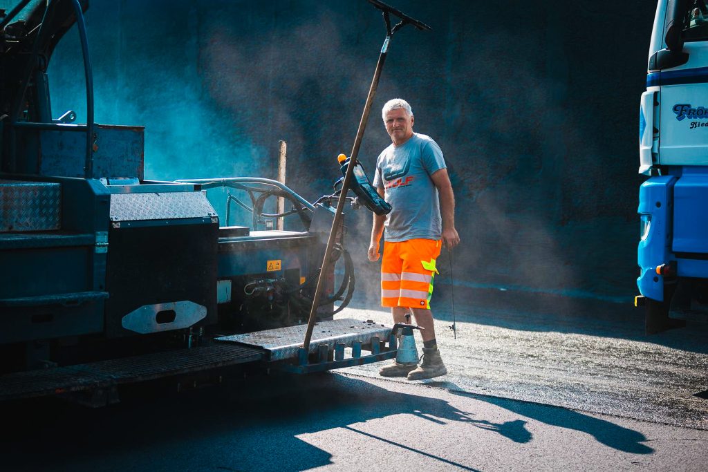 Ein Mitarbeiter lächelt in die Kamera, während er neben einem  Asphaltfertiger steht.