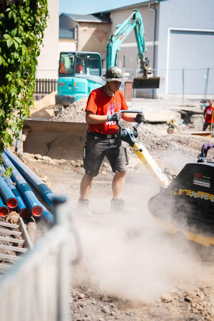 Ein Arrbeiter, wie er eine Rüttelplatte auf einer Baustelle bedient. Staub wirbelt auf, im Hintergrund sind weitere Baumaschinen und Rohre zu erkennen.