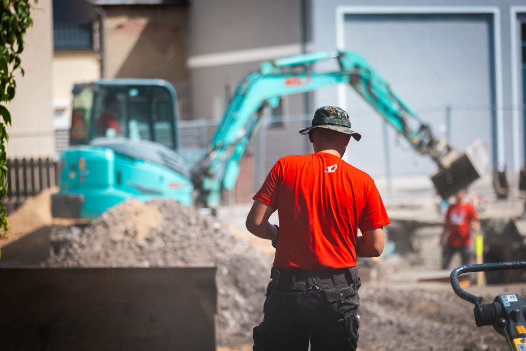 Rückenansicht eines Bauarbeiters, der eine Baustelle überwacht. Im Hintergrund sind ein Bagger und weitere Kollegen bei der Arbeit zu sehen.