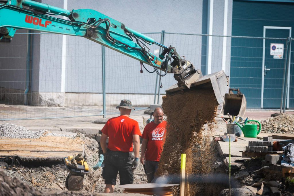 Zwei Bauarbeiter in roten T-Shirts der Firma Wolf Straßen- und Tiefbau bei Erdarbeiten. Ein Bagger mit dem Schriftzug WOLF schüttet Erde in eine Grube, während die Arbeiter die Baustelle überwachen.