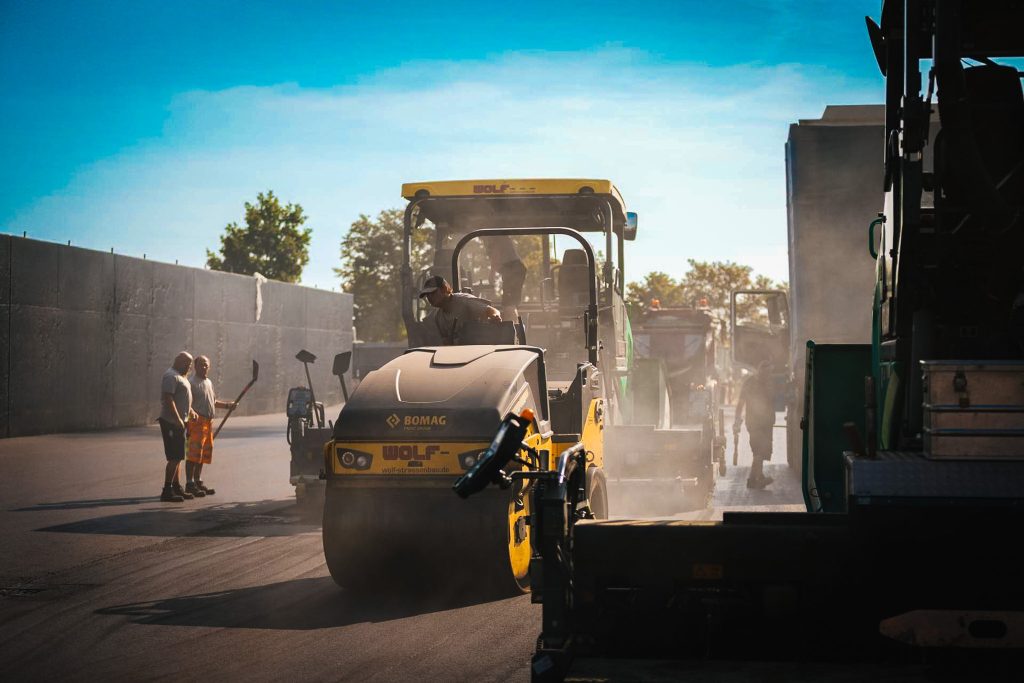Straßenwalze (BOMAG) beim Asphaltieren einer Straße mit Bauarbeitern und Staubentwicklung unter blauem Himmel.