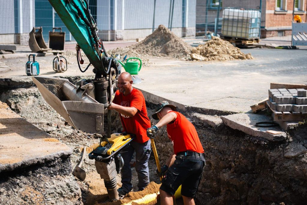 Zwei fröhliche Mitarbeiter bei der Koordination an einer Baustelle. Einer der Arbeiter bedient eine Rüttelplatte, während sein Kollege im Hintergrund eine Schaufel hält. Ein Bagger wird genutzt, um das Material präzise zu verarbeiten.