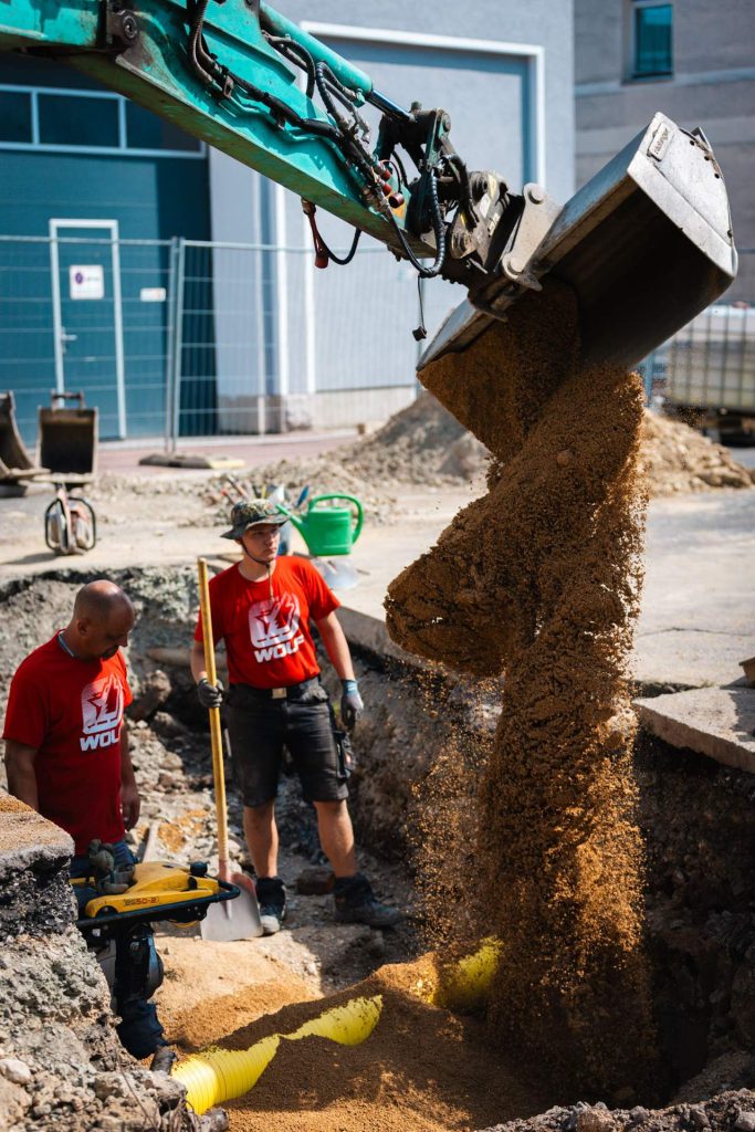 Detailaufnahme, die zeigt, wie ein Bagger Erde in eine Grube schüttet, während zwei Mitarbeiter danebenstehen und die Arbeiten überwachen.