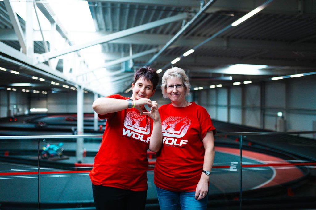 Zwei Mitarbeiterinnen im Wolf-Shirt vor der Kartbahn, eine zeigt mit den Händen ein Herz
