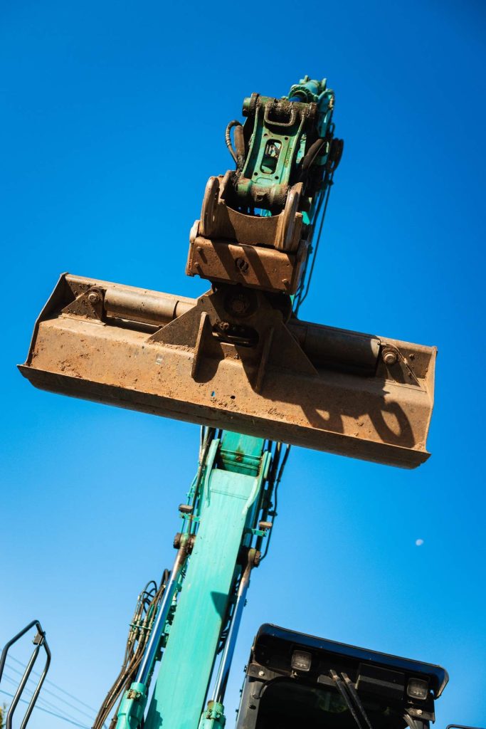 Nahaufnahme einer robusten, bräunlichen Baggerschaufel, die bei den Erdarbeiten auf der Baustelle zum Einsatz kommt. Die Schaufel ragt in den klaren, blauen Himmel.