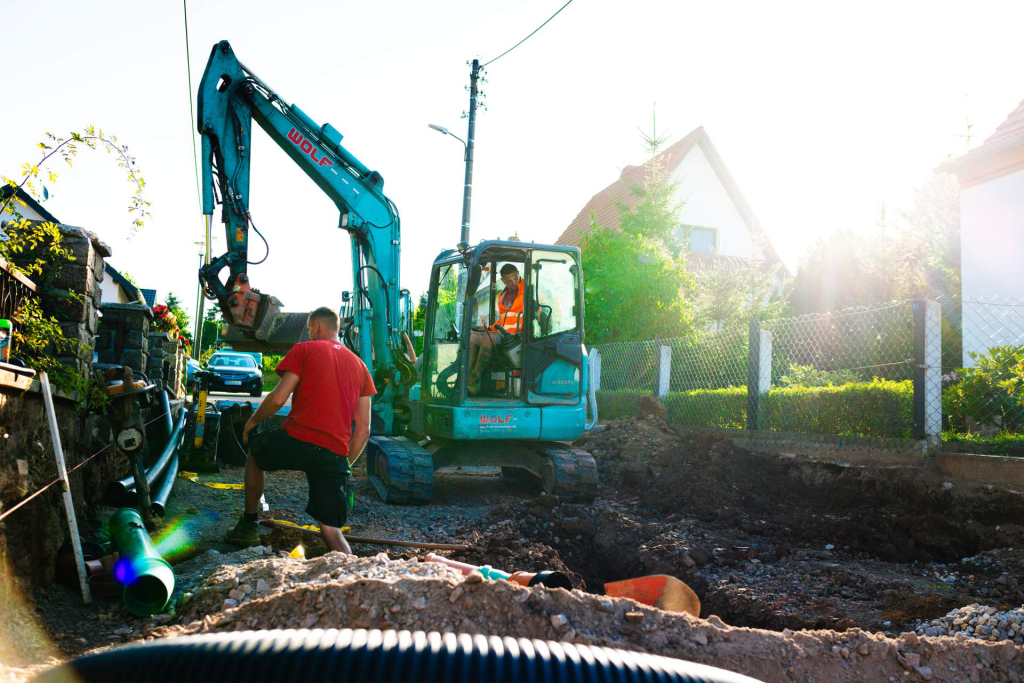 Zwei Bauarbeiter der Firma Wolf sind an einem Graben auf der Baustelle in der Siedlerstraße in Friedrichsgrün beschäftigt. Einer bedient einen Bagger, der Erde bewegt, während der andere neben dem Graben steht. Die Sonne scheint hinter Häusern hervor