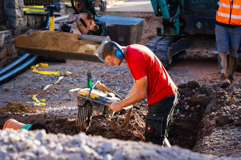 Ein Bauarbeiter der Firma Wolf, der einen Gehörschutz trägt, bedient einen Stampfer, um den Boden in einem Graben zu verdichten. Er befindet sich mitten im Geschehen auf der Baustelle.
