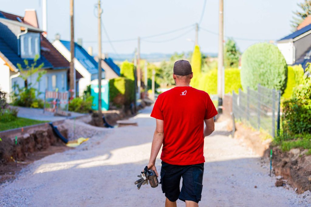 Ein Bauarbeiter der Firma Wolf, von hinten fotografiert, geht mit Handschuhen in der Hand Siedlerstraße entlang. Die Straße ist auf beiden Seiten von Gräben und Häusern gesäumt.