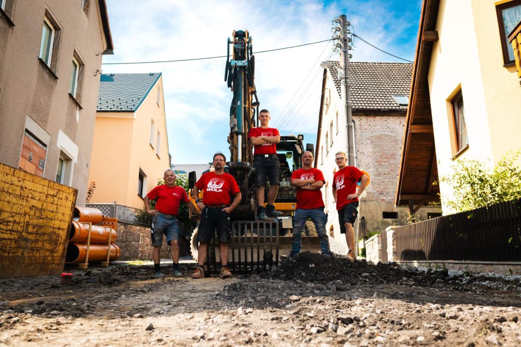 Das gesamte Team der Firma Wolf Straßen- und Tiefbau posiert gemeinsam auf der Baustelle. Die Bauarbeiter tragen ihre roten Arbeits-T-Shirts und stehen selbstbewusst vor einem großen Bagger.