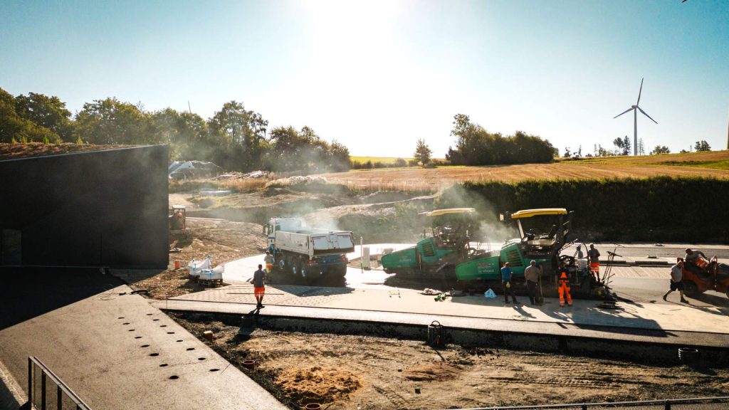 Straßenbau und Asphaltierung eines Vorplatzes oder Parkplatzes. Zwei Vögele Fertiger und ein Kipplaster sind im Einsatz, umgeben von aufsteigendem Dampf. Im Hintergrund ist ein modernes Gebäude und ein Windrad sichtbar.