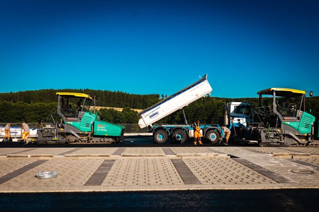 Asphaltierungsarbeiten auf einer Baustelle mit zwei Vögele Straßenfertigern und einem Kippsattelauflieger, der Asphalt abkippt. Bauarbeiter in orangefarbener Schutzkleidung sind beteiligt.