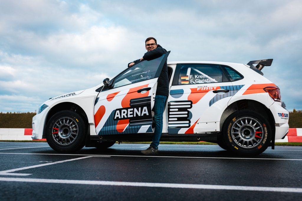 Fabian Kreim lehnt lächelnd an einem weißen VW Polo Rallyeauto auf einer Rennstrecke, Wolkenhimmel.