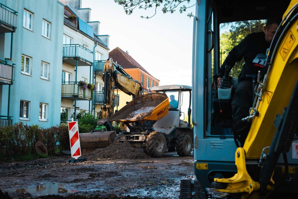 Baustellenaussicht mit einem großen gelben Bagger/Dumper, der Schmutz vor einem Wohnhaus lädt, mit der Kabine eines kleineren Baggers im Vordergrund.