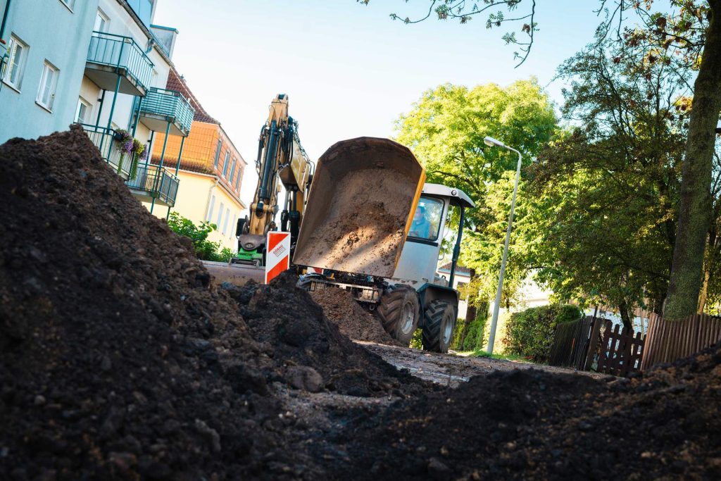 Blick über einen großen Haufen dunkler Erde auf einen gelben Bagger/Dumper, der auf einer schlammigen Straße in der Nähe von Wohnhäusern arbeitet.