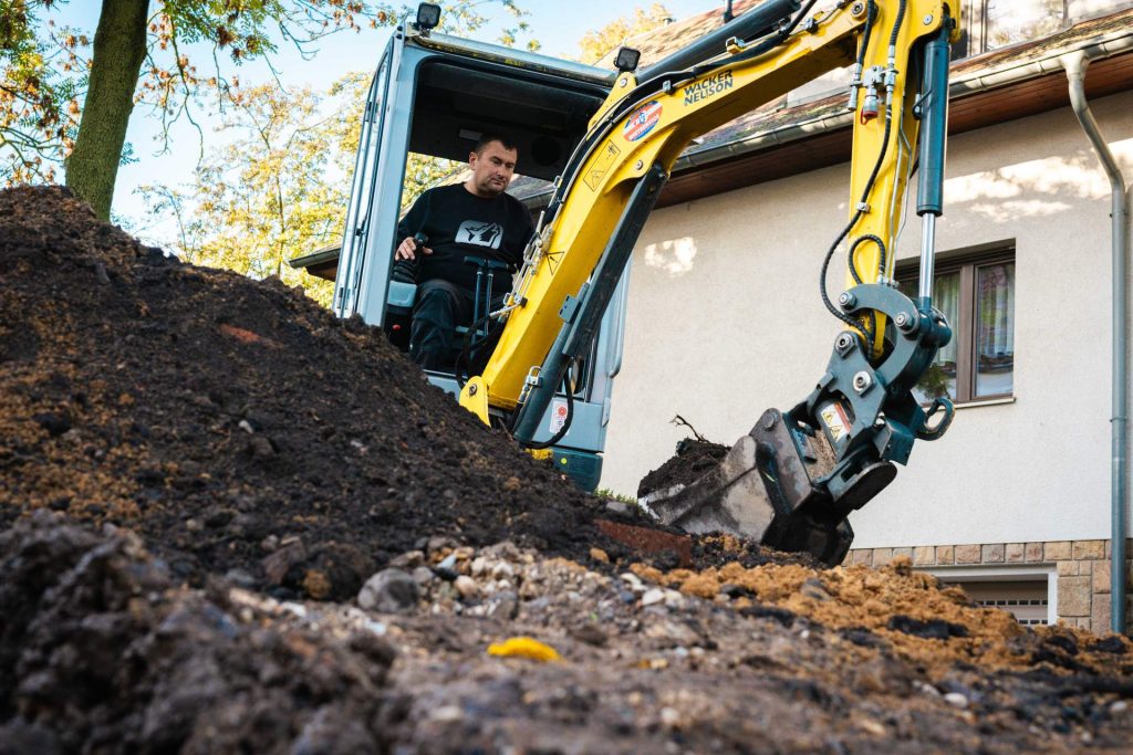 Mann bedient einen gelben Wacker Neuson Bagger und bewegt auf einer Baustelle eine Schaufel voller dunkler Erde und Schmutz.