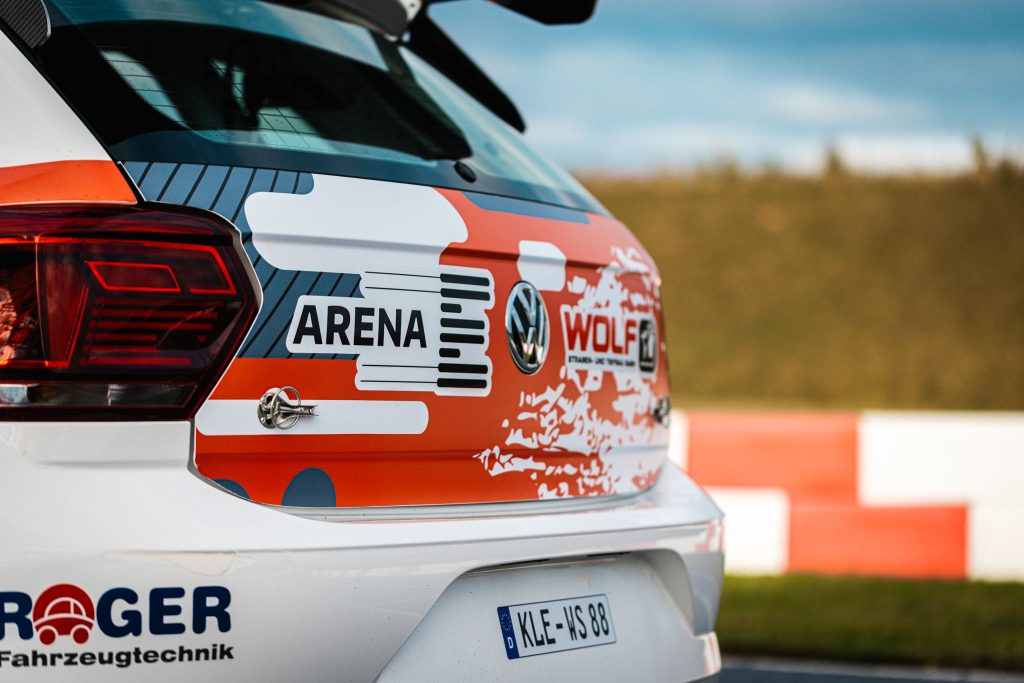 Heckansicht eines weißen VW Polo Rallyeautos mit "ARENA" und "WOLF" Logos und deutschem Kennzeichen auf einer Rennstrecke.