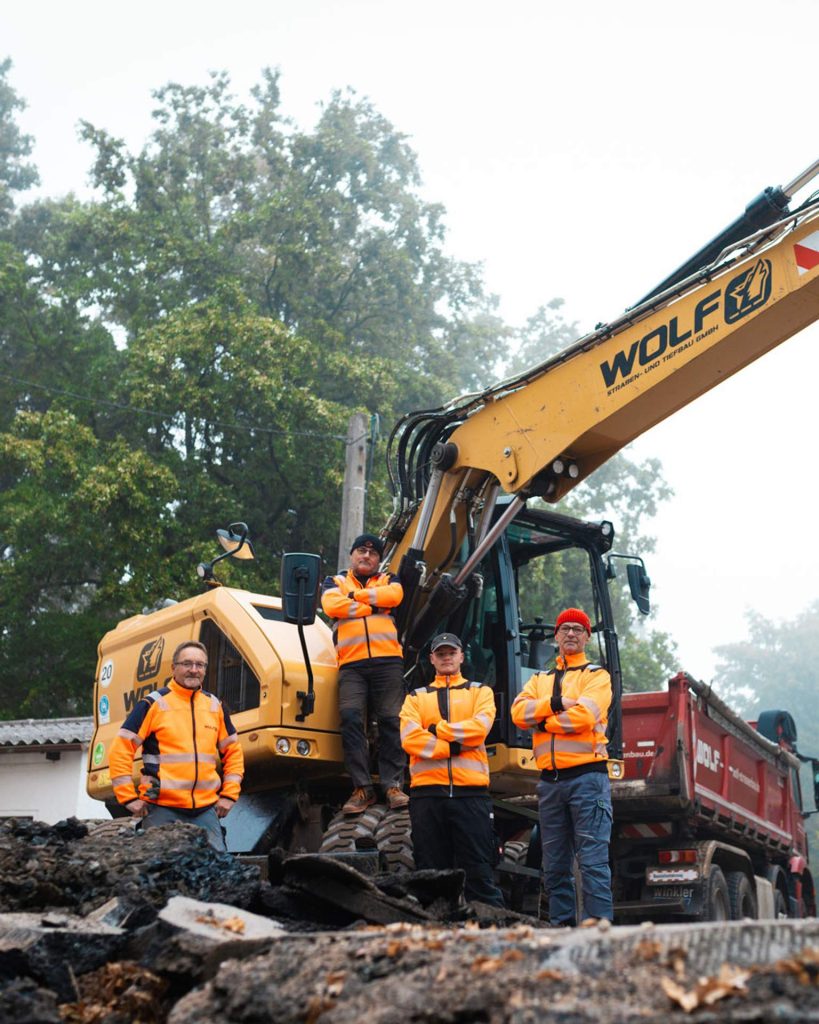 Ein Team von Mitarbeitern der WOLF Straßen- und Tiefbau GmbH posiert in oranger Warnkleidung vor einem gelben Bagger und einem roten LKW auf der Baustelle. Starke Gemeinschaft im Tiefbau.