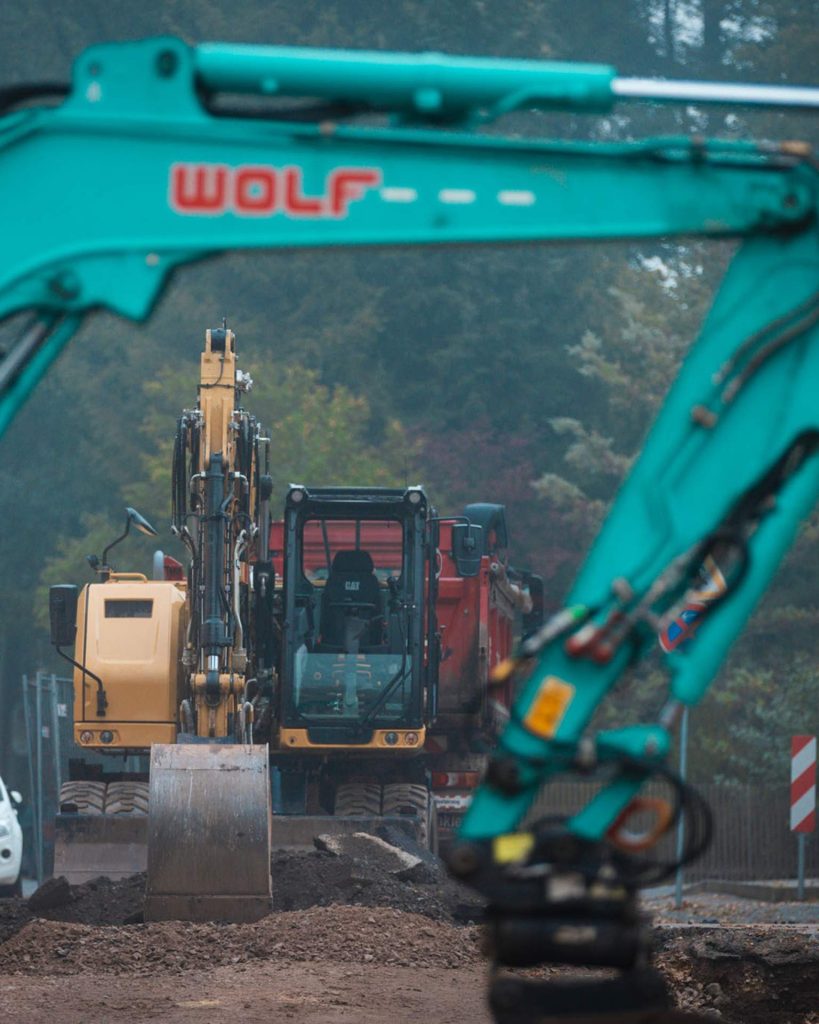 Nahaufnahme eines WOLF Baggers bei der Arbeit, wobei ein türkiser Bagger im Vordergrund steht. Einsatz von moderner Technik der WOLF Straßen- und Tiefbau GmbH auf der Baustelle.