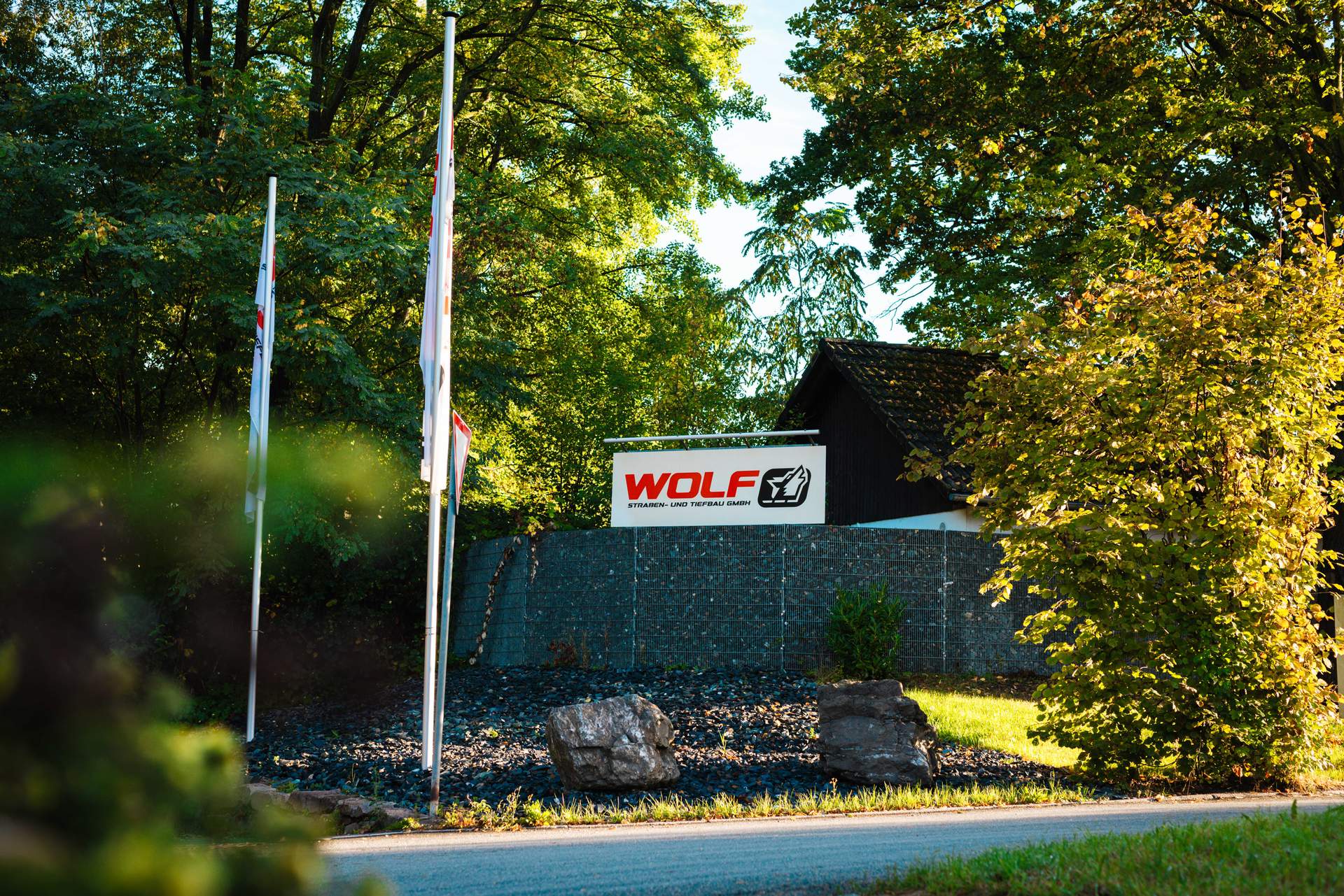 Ein großes slbernes Schild mit dem roten Logo der WOLF Straßen- und Tiefbau GmbH steht eingebettet in eine grüne Bepflanzung. Dies markiert den Firmensitz des erfolgreichen Tiefbau- und Straßenbau-Unternehmens.