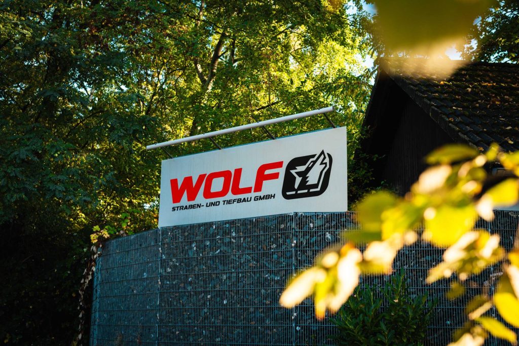 Detailaufnahme des Außenschildes der WOLF Straßen- und Tiefbau GmbH aus der Nähe, umgeben von Hecken. Die klare Präsentation unterstreicht die lokale Präsenz des Tiefbau-Unternehmens.