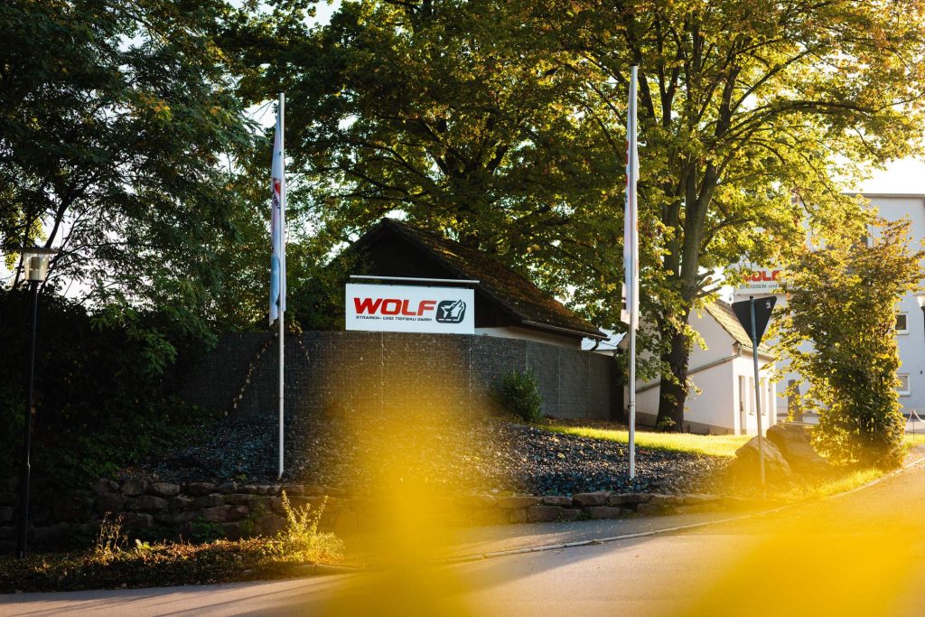 Der Eingangsbereich des WOLF-Hauptsitzes, umrahmt von Bäumen, mit wehenden Flaggen. Das Unternehmen ist ein verlässlicher Partner für Straßenbau- und Tiefbau-Projekte in der Region.