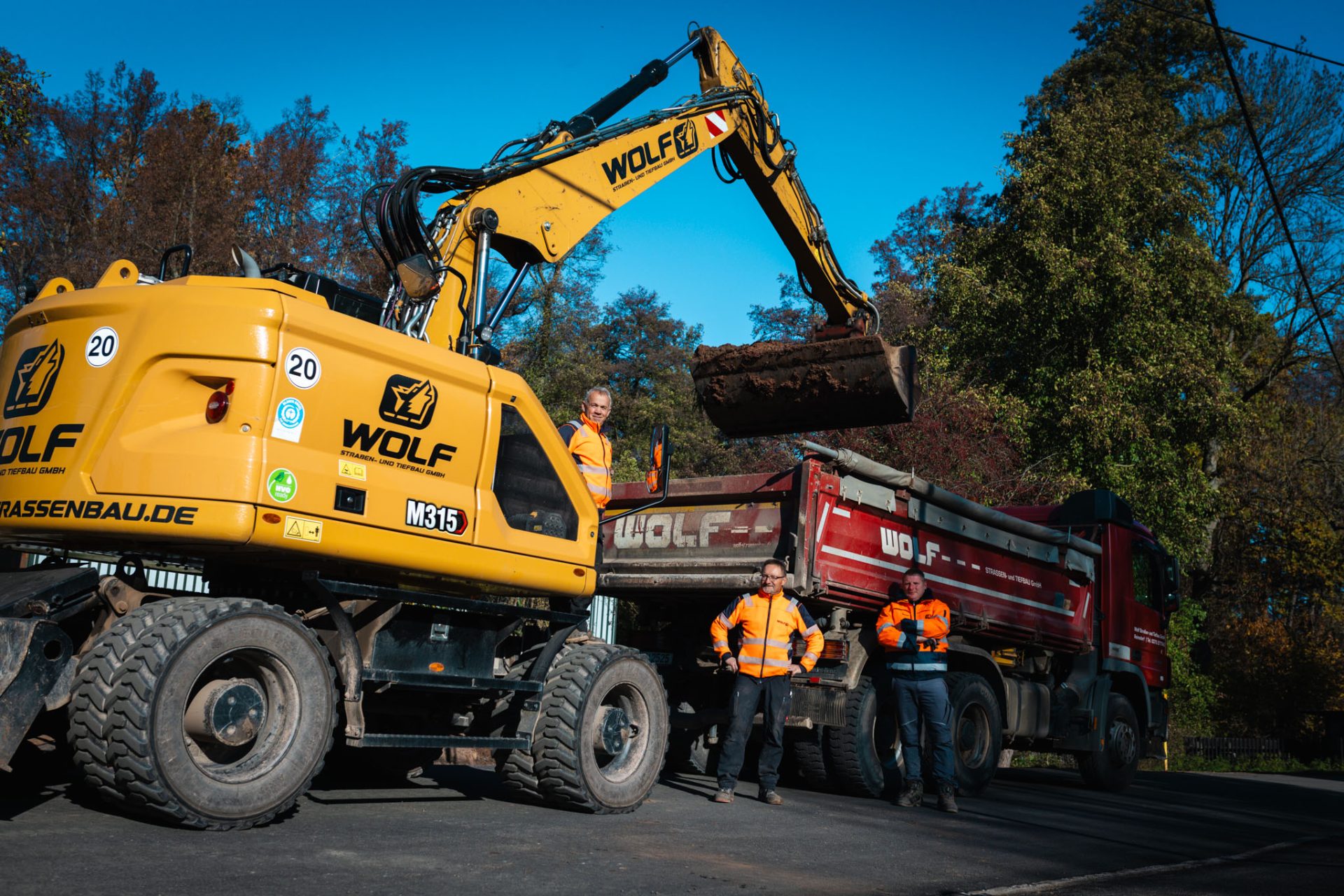 Straßenbauarbeiten bei Sonnenschein: Ein gelber Mobilbagger des Typs Wolf M315 Straßenbau belädt einen roten Wolf Mulden-LKW mit Erde. Drei Bauarbeiter in leuchtend oranger Arbeitskleidung stehen lächelnd neben den großen Baufahrzeugen auf der Baustelle.
