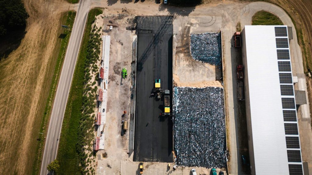 Drohnenaufnahme einer industriellen Baustelle mit Asphaltierungsarbeiten neben einer großen Lagerhalle mit Photovoltaikanlage.