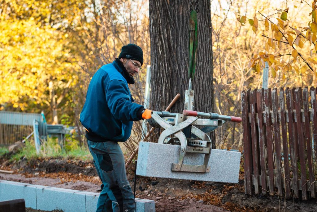 Einsatz einer Bordsteinzange am Baggerarm: Mechanische Unterstützung beim Versetzen schwerer Betonelemente für die Straßenentwässerung und Randbefestigung.