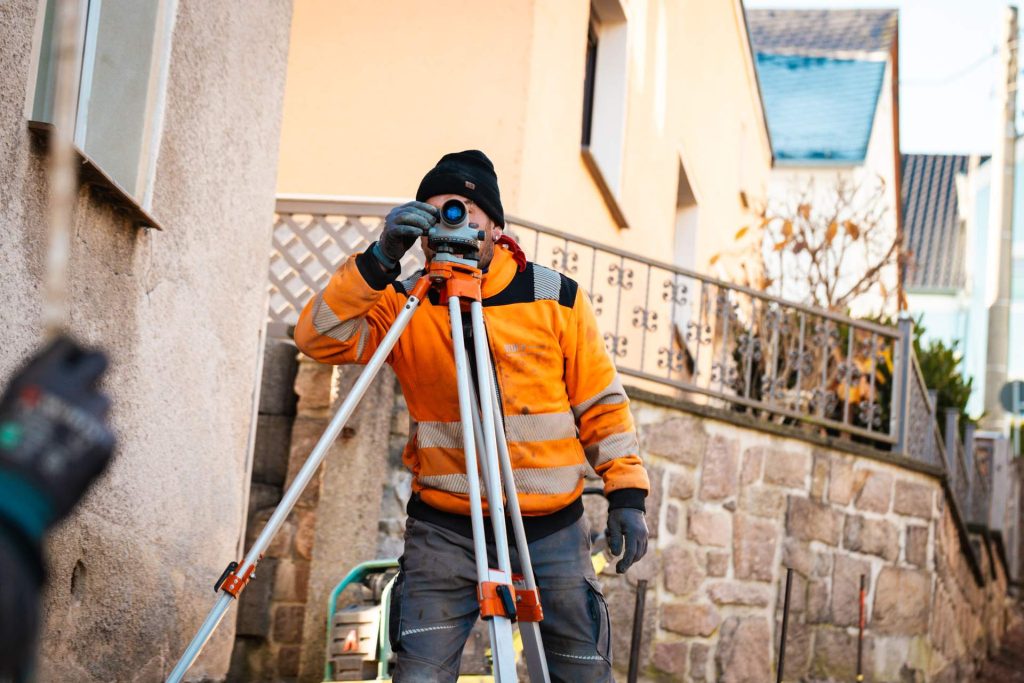 Bauarbeiter in orangefarbener Warnkleidung bedient ein Nivelliergerät auf einem Stativ zur präzisen Höhenmessung auf einer Baustelle.