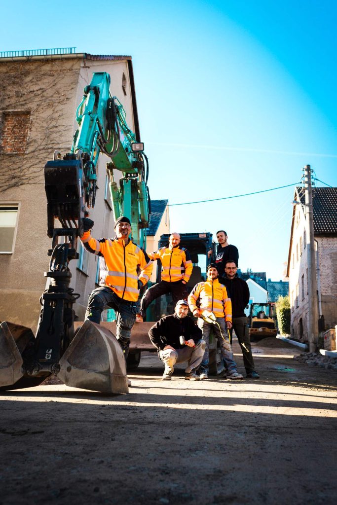 Stolzes Team von Bauarbeitern posiert vor einem türkisfarbenen Bagger auf einer fertiggestellten oder laufenden Baustelle.