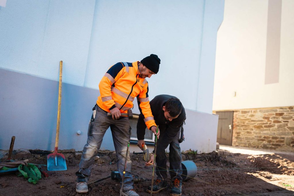 Zwei Bauarbeiter in Arbeitskleidung arbeiten gemeinsam am Boden an der Ausrichtung von Schnurgerüsten für die Pflasterung.