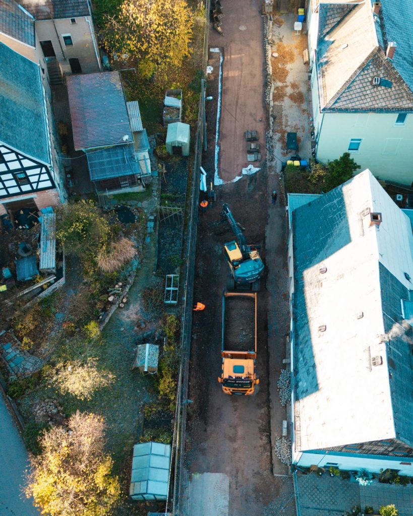 Logistik und Maschineneinsatz in einer engen Wohnstraße in Wildenfels. LKW und Bagger arbeiten Hand in Hand, um den alten Belag abzutransportieren und Platz für den neuen Straßenaufbau zu schaffen.