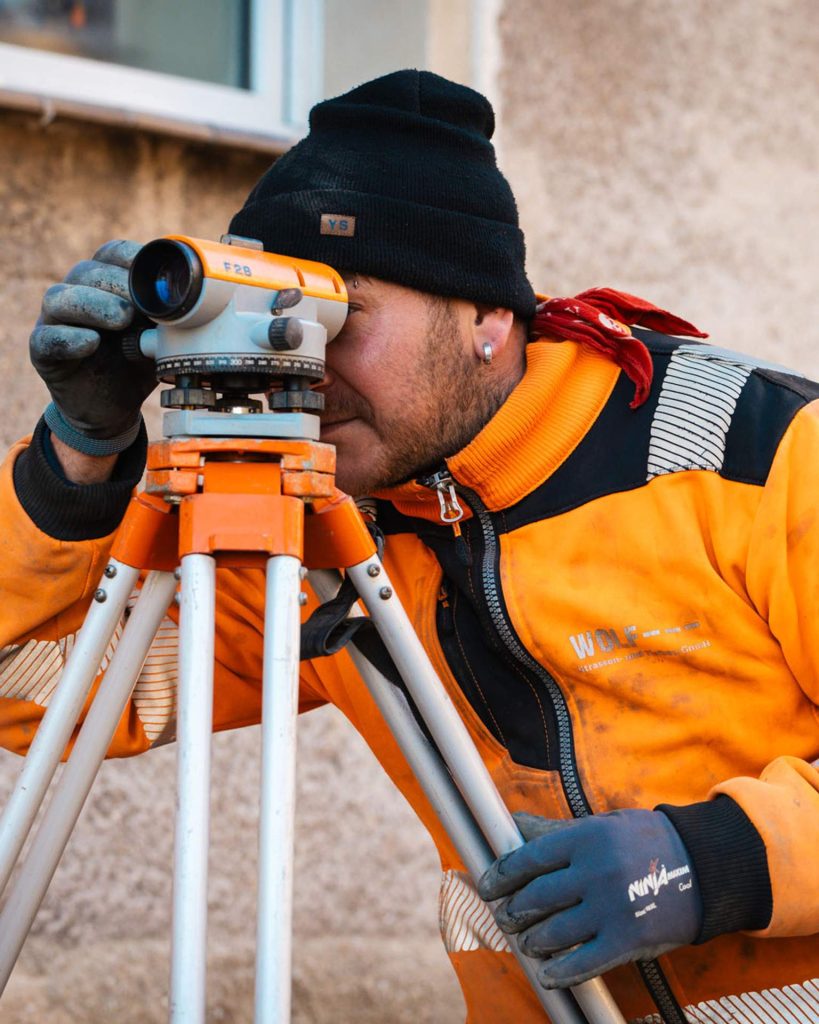 Detailaufnahme eines Vermessungstechnikers mit Mütze beim Blick durch ein optisches Nivelliergerät auf einer Außenbaustelle.