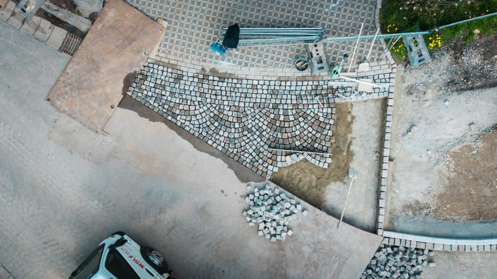 Detailaufnahme aus der Vogelperspektive auf einen Bereich, in dem Kopfsteinpflaster in einem fächerförmigen Muster neu verlegt wird. Daneben liegen Werkzeuge wie eine Schaufel, eine Gießkanne und ein Stapel Pflastersteine.