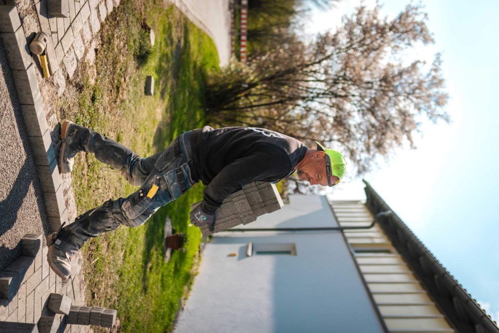 Profilansicht eines Mitarbeiters, der mehrere Pflastersteine zur Verlegestelle trägt. Im Hintergrund blühen Bäume, was den frühlingshaften Fortschritt der Baustelle unterstreicht.